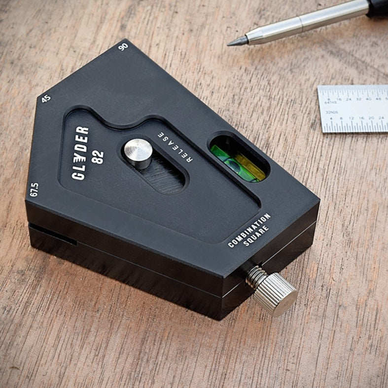 A black GLYDER 82 combination square tool made of T6 aluminum, with a stainless steel thumb screw and a built-in bubble level, placed on a wooden surface next to a pen and a smaller ruler.